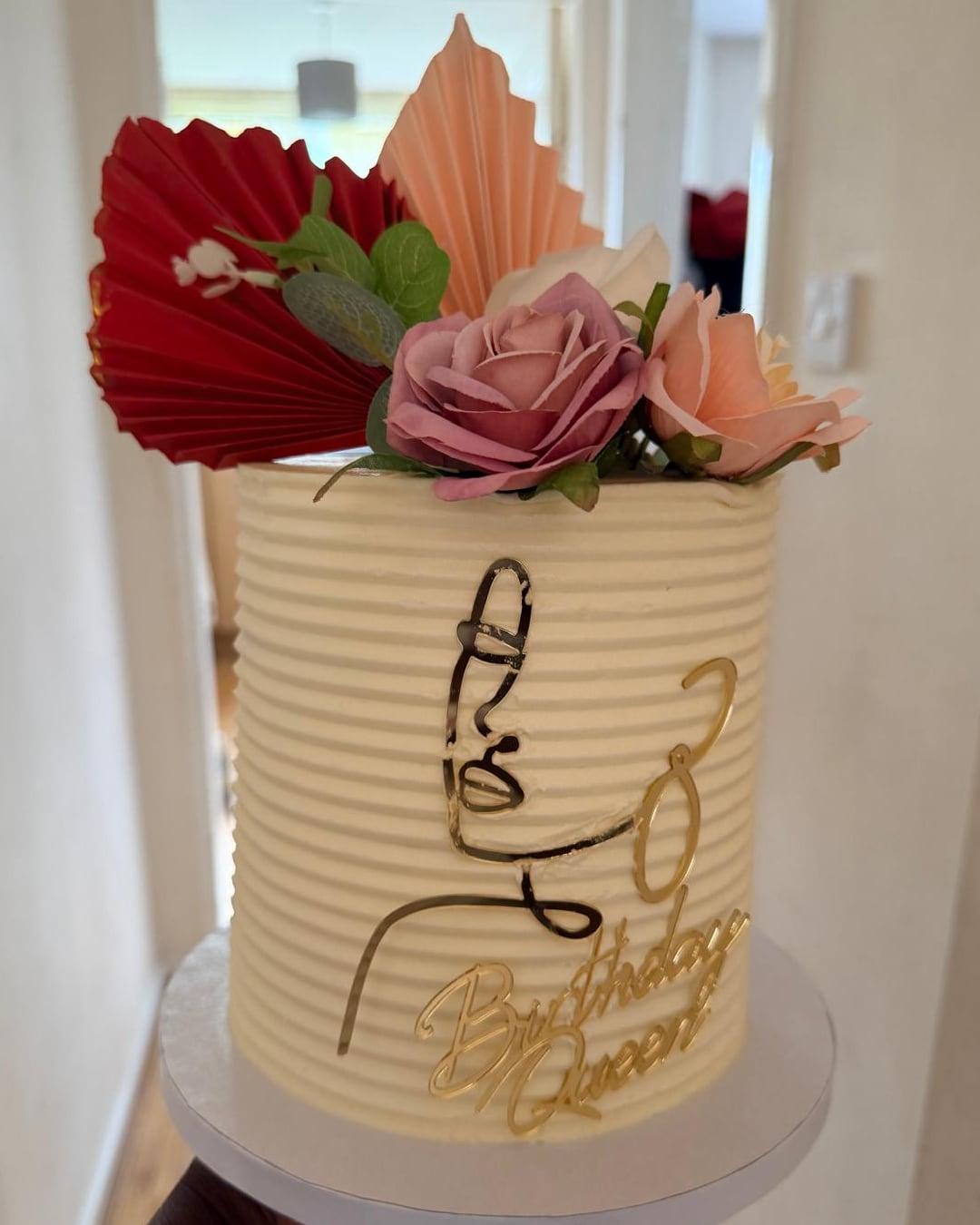 A cream ribbed buttercream cake with a gold script drawing and paper florals