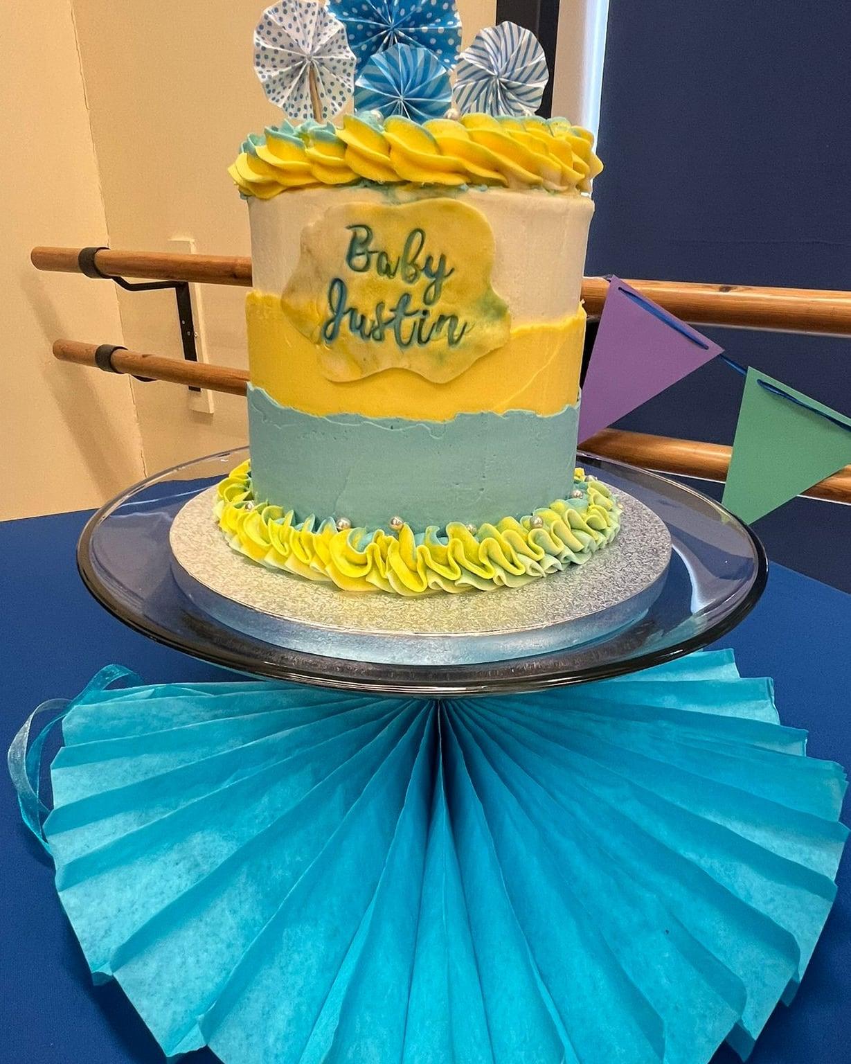 A yellow and sage baby celebration cake with a hand-piped shell border and a named plaque