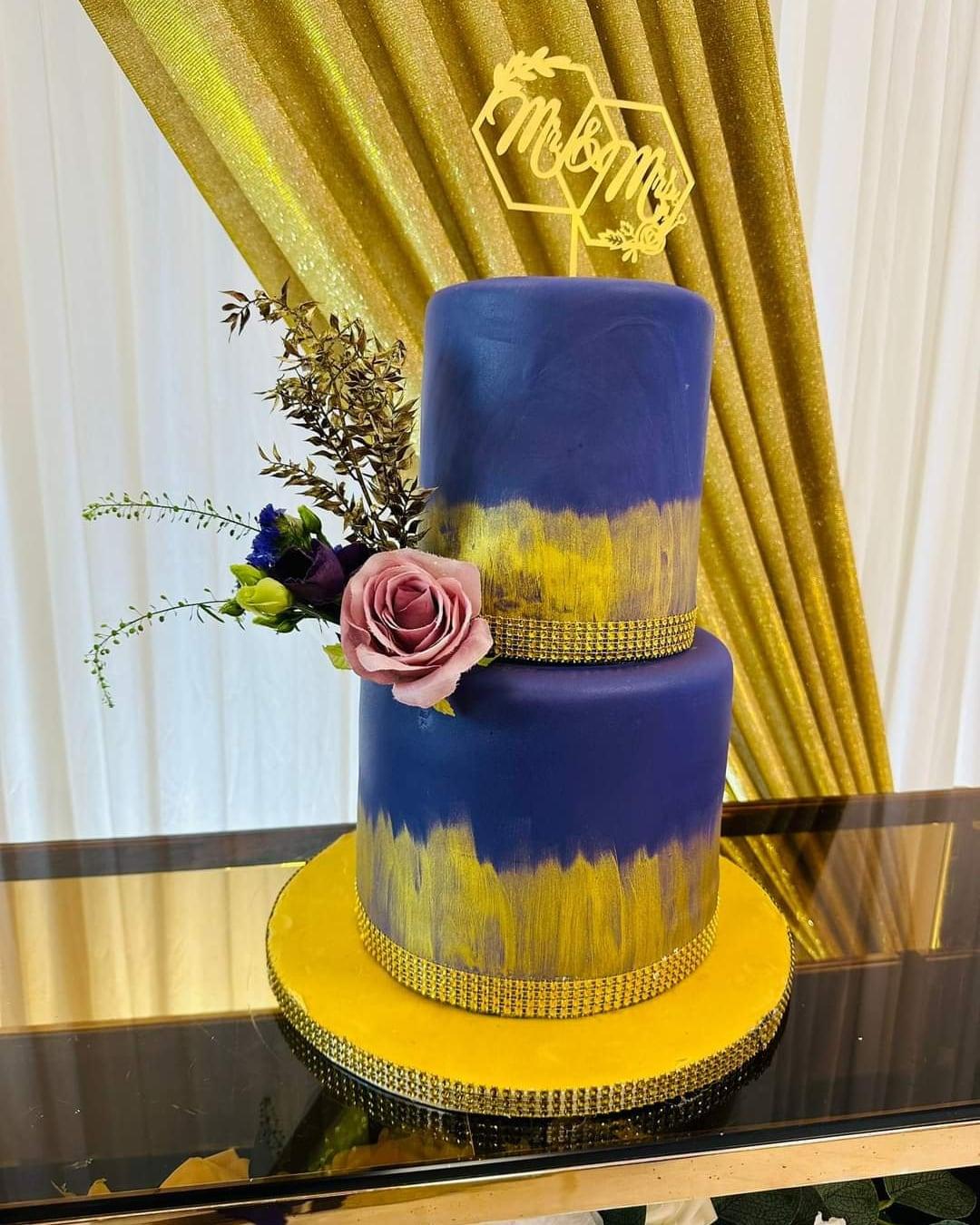 A navy and gold two-tier wedding cake dressed with a sugar rose, photographed against gold drapery
