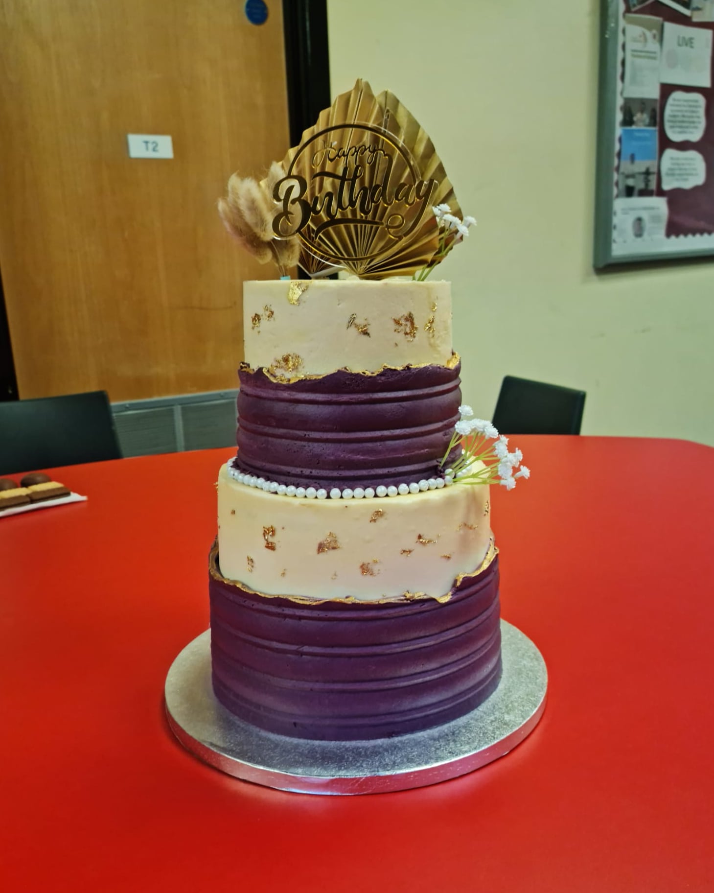 A three-tier plum and ivory celebration cake finished with gold leaf, piped pearls and a gold fan topper